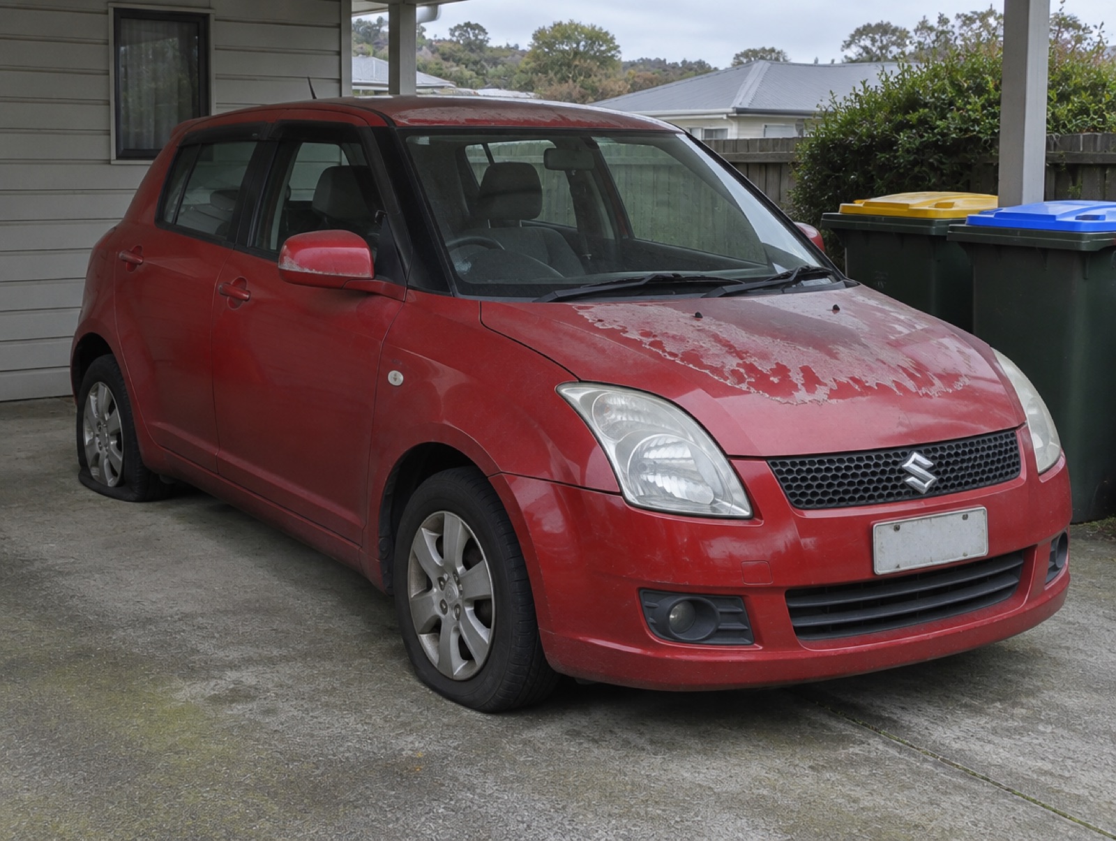 A tired red hatchback with peeling paint and a flat tyre in a carport