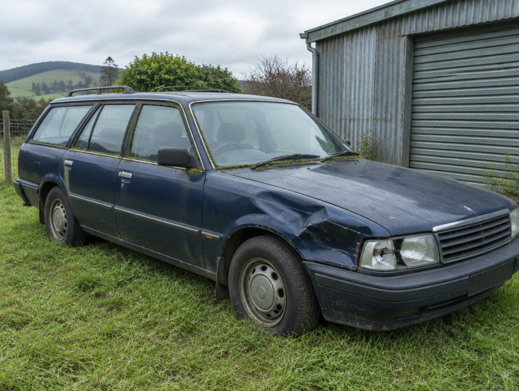 An old dark blue station wagon with a dented fender beside a corrugated iron garage
